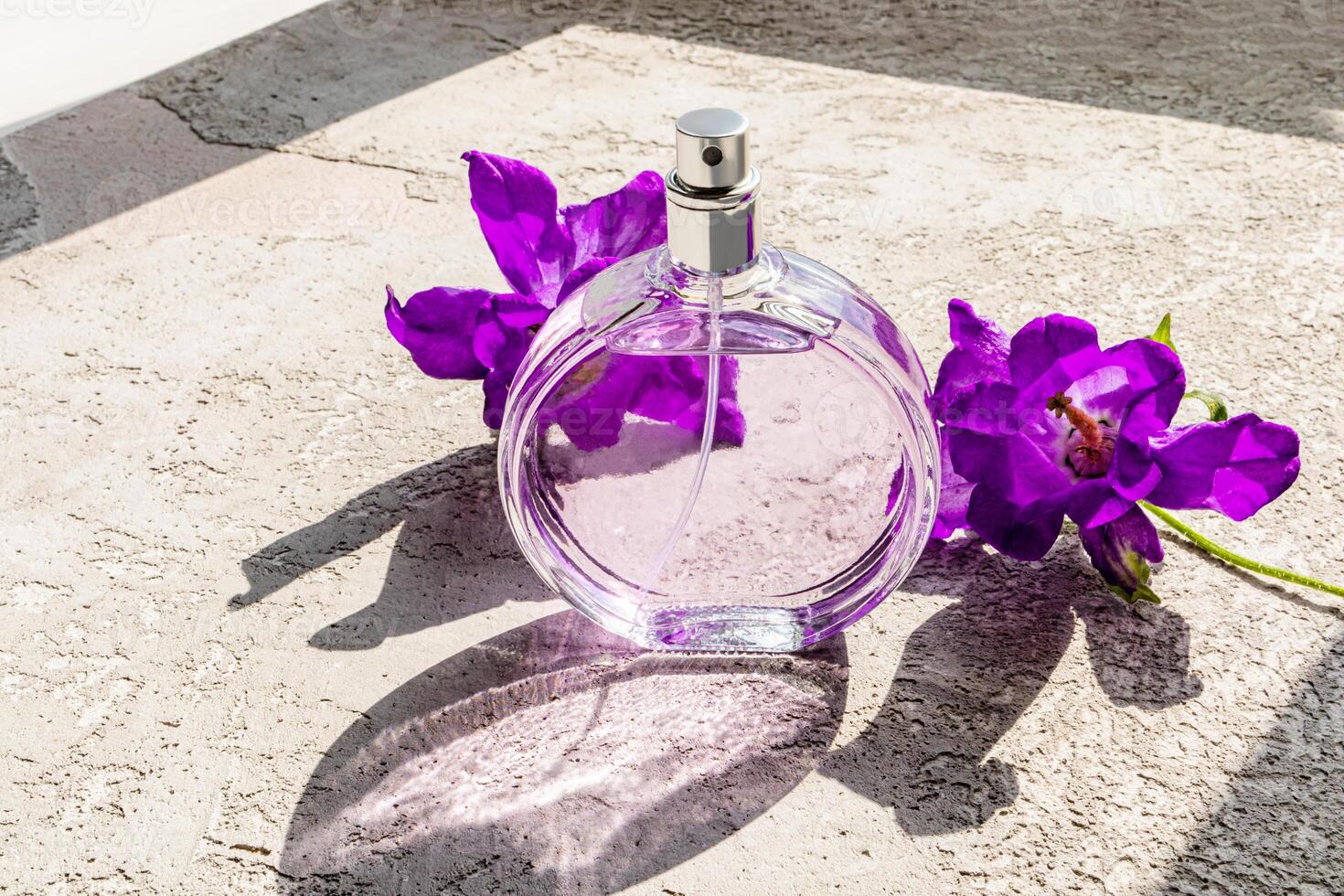 A beautiful bottle of women's perfumes stands on a concrete surface with lilac iris flowers in the sun. A blank bottle mockup. Product presentation. photo