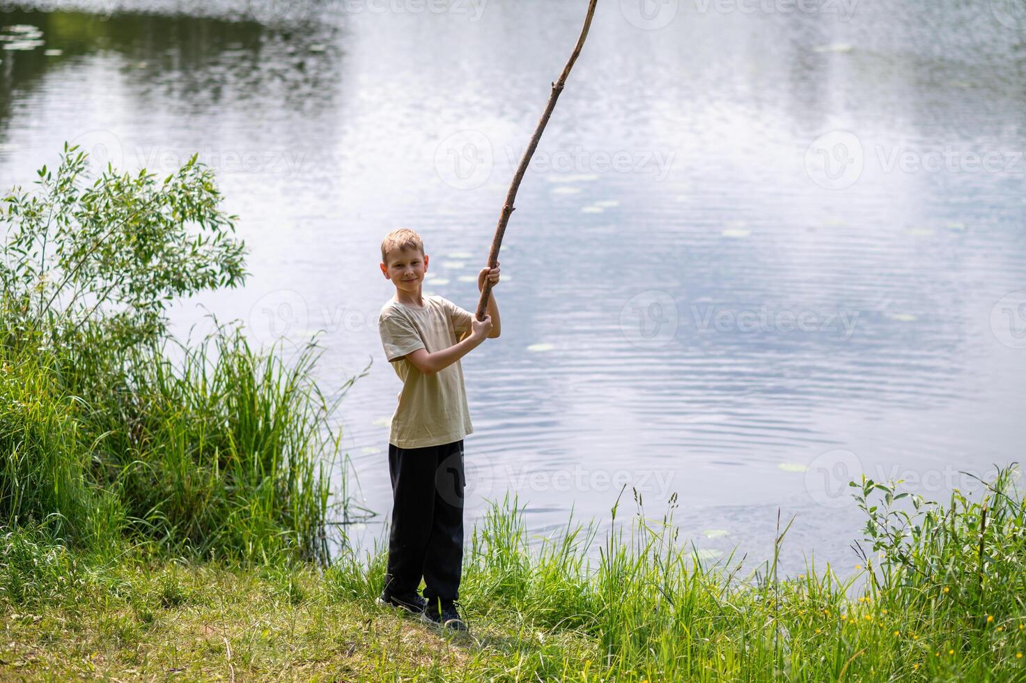 Boy standing on lakeside with a long stick, playing near the water in nature on a summer day photo