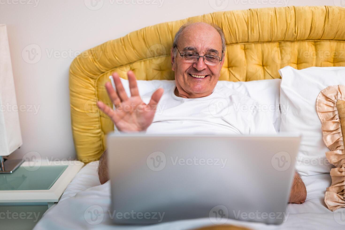 Senior man in pajamas sitting on bed, smiling and waving during call on his laptop, enjoying remote work from home. photo
