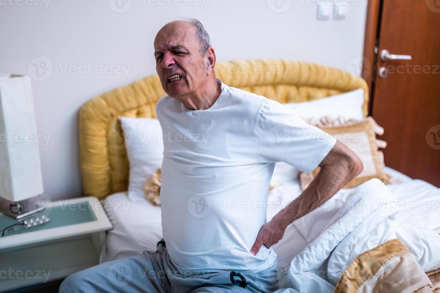 Senior man sitting on the edge of the bed in the morning, clutching his back in pain due to a backache and arthritis. photo