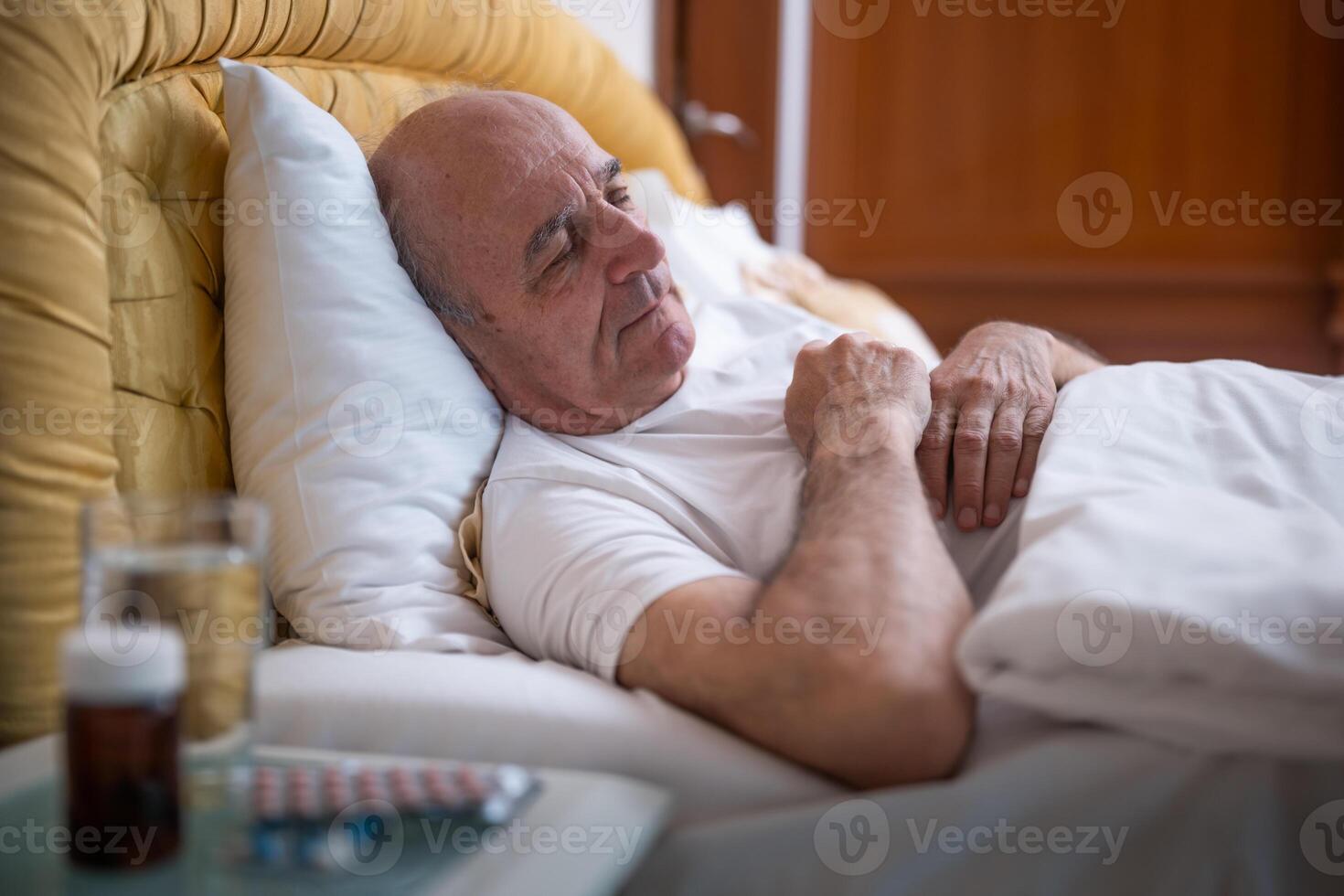 Elderly Caucasian male with gray hair sleeping in bedroom, holding a glass of water and capsule for health care on night table next to his bed. photo