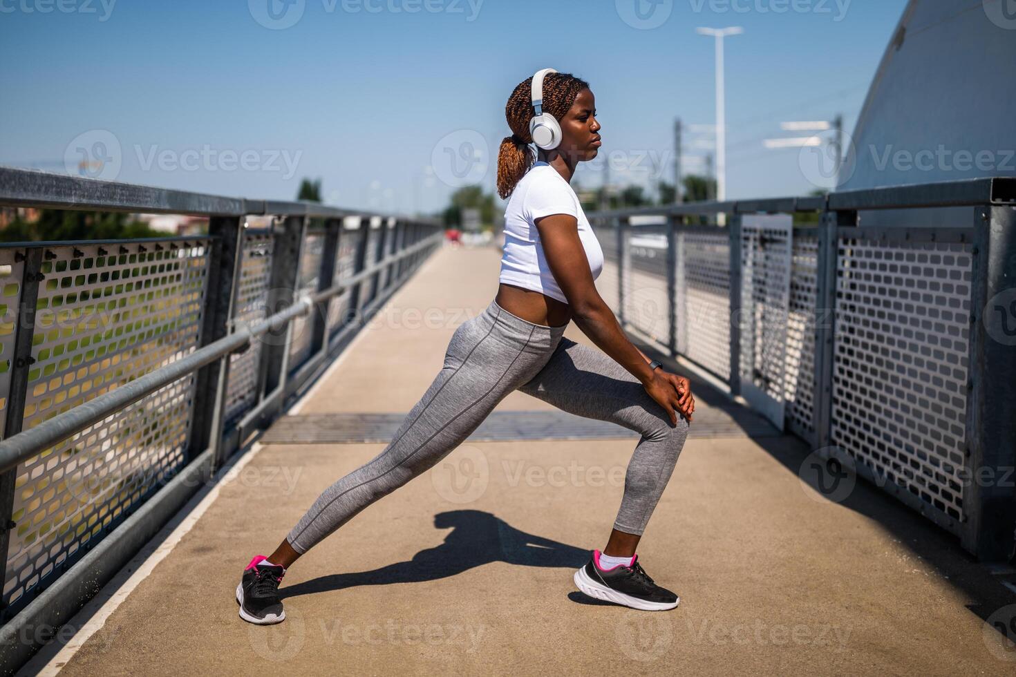 Motivated young African-American woman in sportswear with headphones enjoying exercising and stretching while standing at the city bridge. She is in the squatting position. photo
