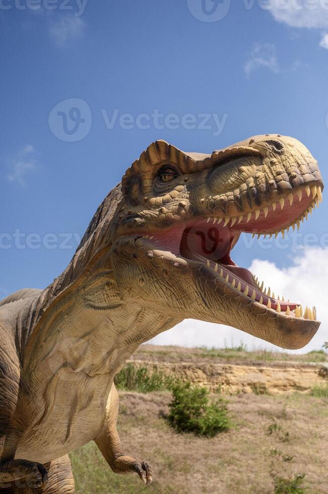 Huge Artificial dinosaur tyrannosaurus, close-up of dinosaur head against sky background. With copy space. Dinosaur has open mouth, teeth, front legs are visible photo
