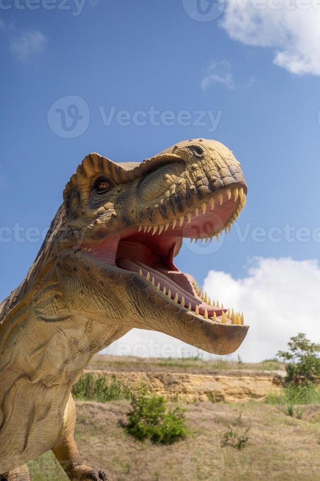 Huge Artificial dinosaur tyrannosaurus, close-up of dinosaur head against sky background. With copy space. Dinosaur has open mouth, you can see its teeth, front legs. Yellow rocks in the background photo