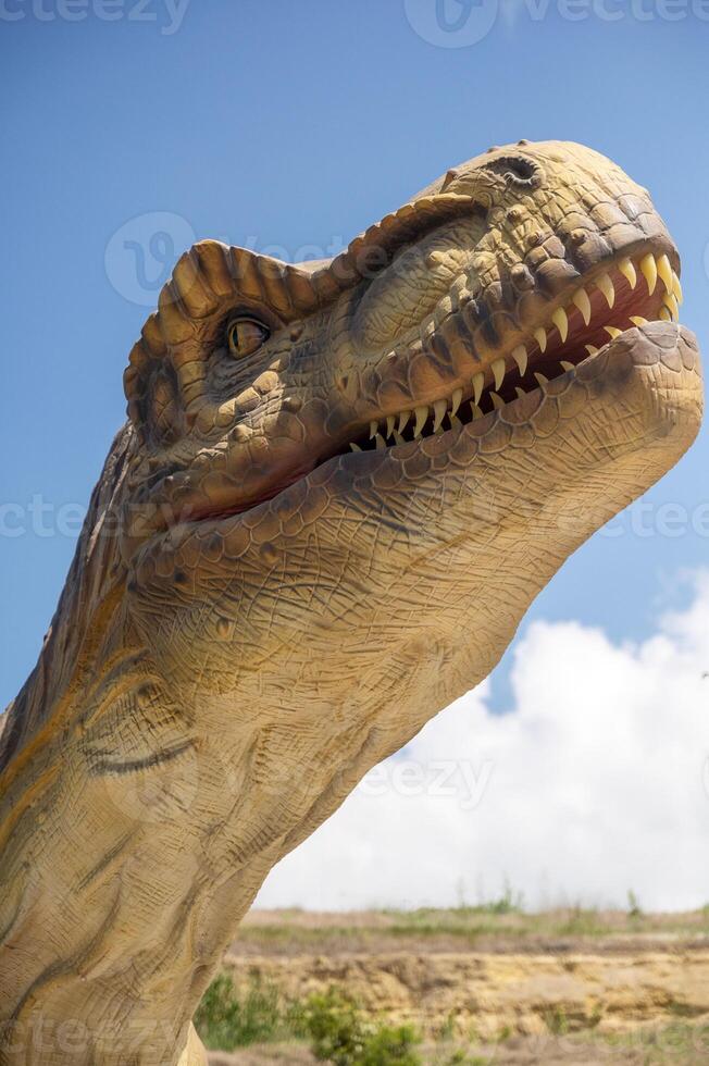 Huge Artificial dinosaur tyrannosaurus, close-up of dinosaur head against sky. With copy space. Dinosaur shows its teeth, yellow rocks and trees in the background photo