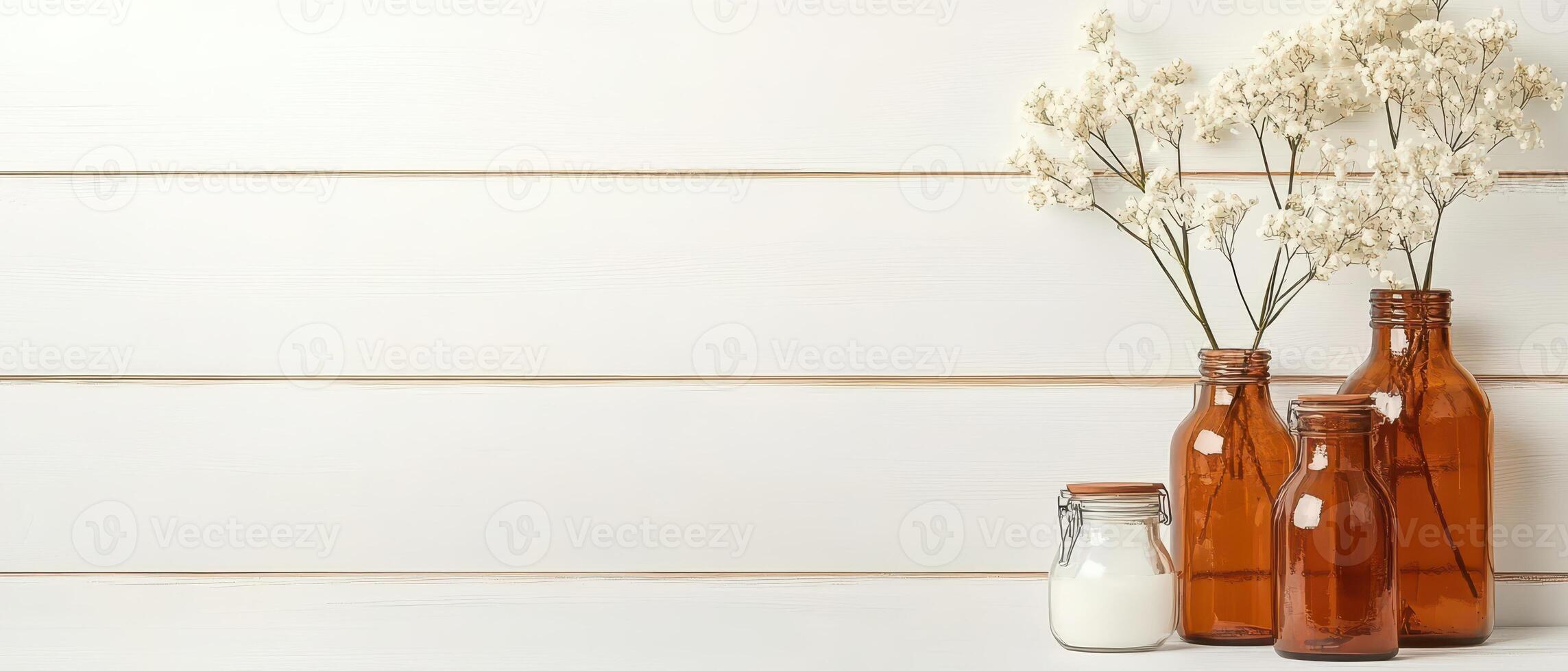 Dried white flowers displayed in brown glass bottles near a full glass jar on a white wooden surface against a wall photo