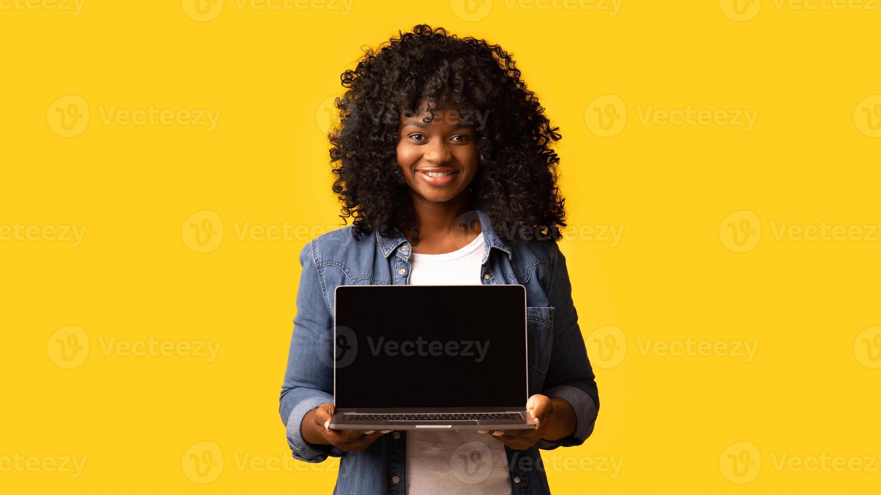 Cheerful beautiful black woman using laptop with empty screen over yellow studio background, copy space. Cute african american young lady showing notebook with blank display, mockup photo