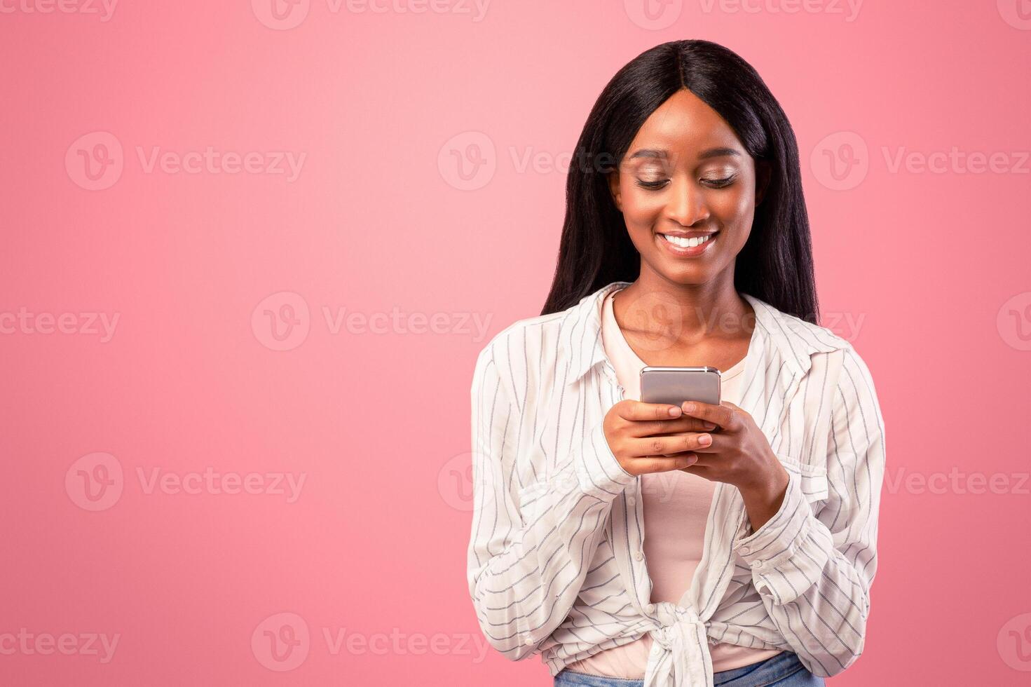 Online life concept. Attractive black woman using smartphone on pink studio background. Pretty African American lady browsing web, sending message, chatting with friend, checking social media photo