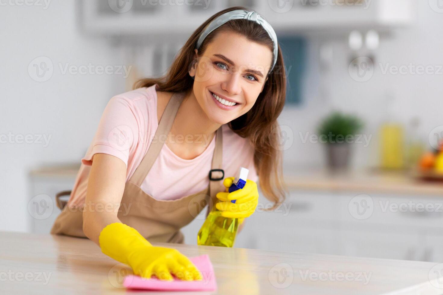 Happy young housewife or maid cleaning table with rag and detergent at kitchen, copy space. Positive millennial lady doing house cleanup. Professional sanitary service concept photo
