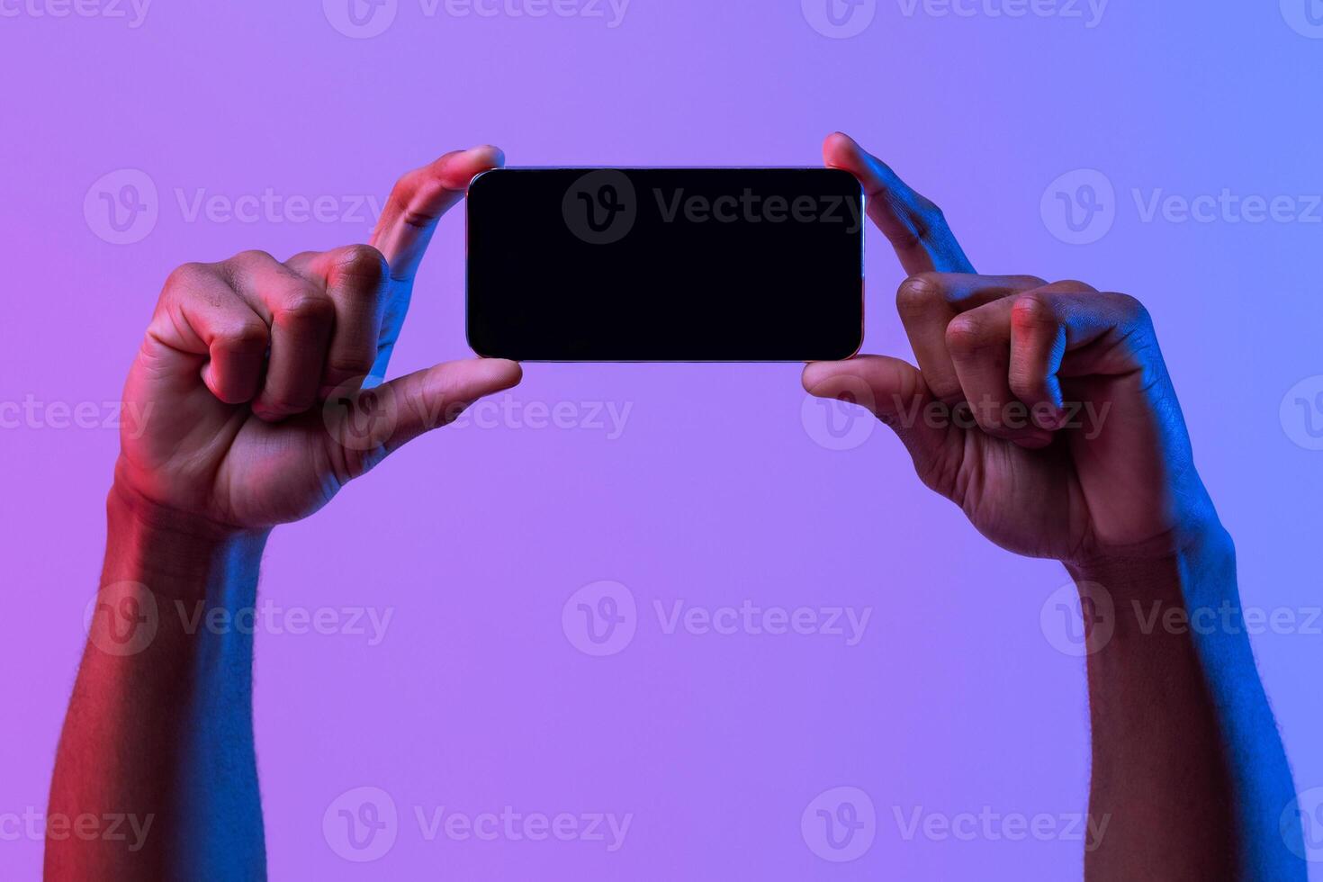 Black Male Hands Holding Smartphone With Black Screen In Horizontal Orientation Under Vivid Neon Light Over Purple Background, Mockup Image With Copy Space For Your App, Game Or Website Design photo