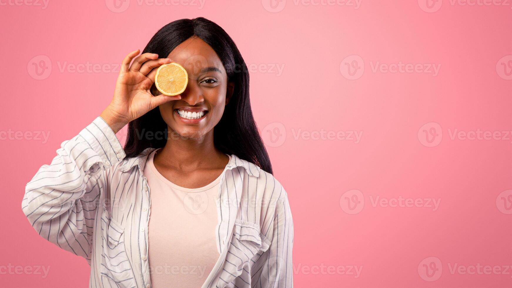 Vitamin C for skincare concept. Beautiful black woman closing her eye with lemon half on pink studio background. African American lady holding citrus fruit in front of her face photo