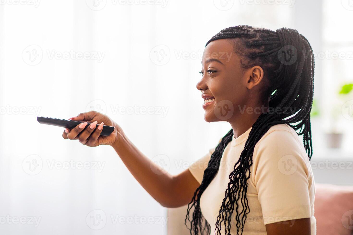 Side view of cheerful african american lady switching channels, home entertainment concept. Happy young black woman sitting on sofa with TV remote, choosing movie and eating popcorn, copy space photo
