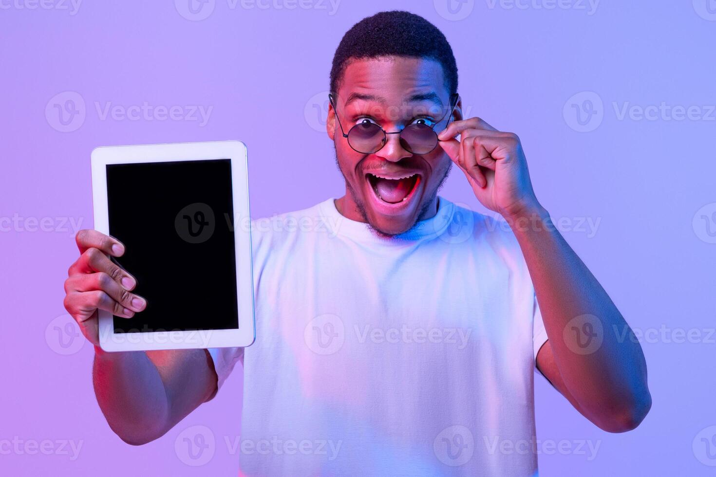 Check This App. Surprised African Guy In Sunglasses Holding Digital Tablet With Black Blank Screen For Mockup, Demonstrating Copy Space For Your Design, Standing In Neon Light On Purple Background photo