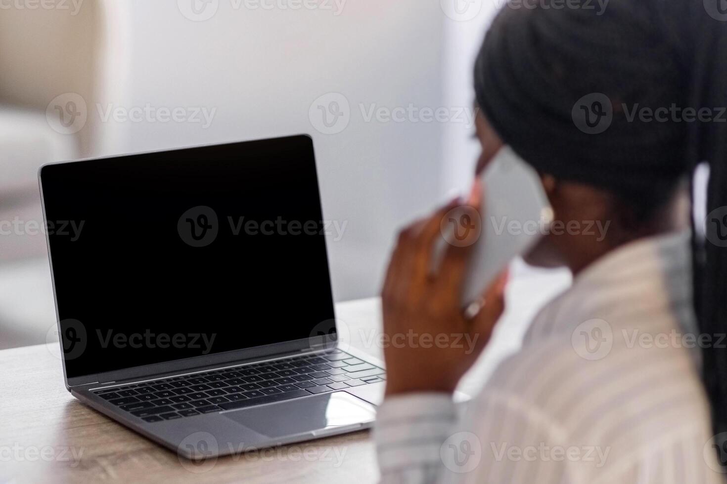 Unrecognizable black woman having conversation on mobile phone, using laptop with blank screen, copy space, back view. African american freelancer talking with clients on smartphone while using pc photo