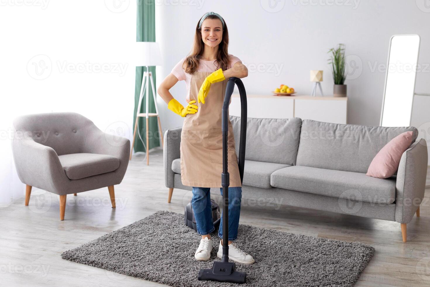 Housecleaning service. Happy young maid with vacuum cleaner standing in middle of tidy living room, full length portrait. Positive millennial housewife doing domestic chores, copy space photo