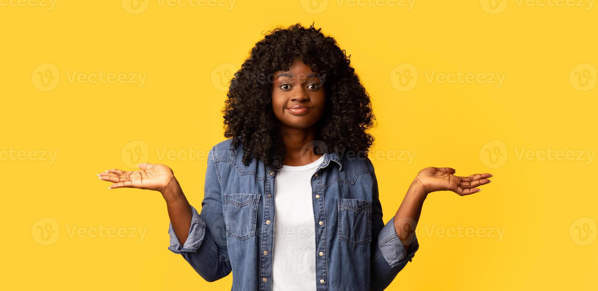 I dont know. Confused young black lady raising hands up and shrugging, yellow studio background, copy space. Pity african american woman with curly hair showing sorrow or compassion photo