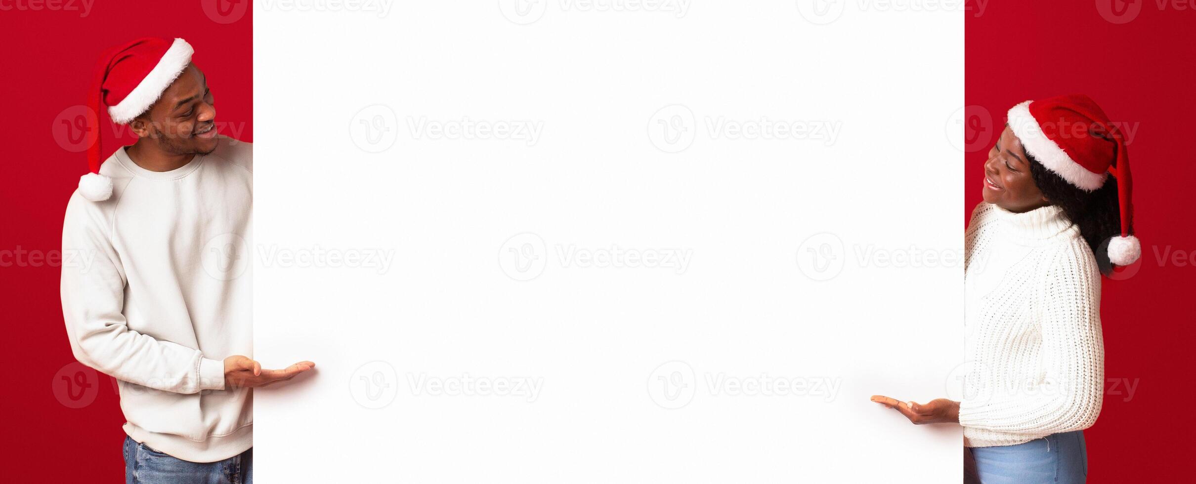 Place For Christmas Ad. Cheerful Black Spouses In Santa Hats Looking And Pointing At White Advertisement Board, Man And Woman Demonstrating Copy Space For Your Text Or Design, Orange Background photo
