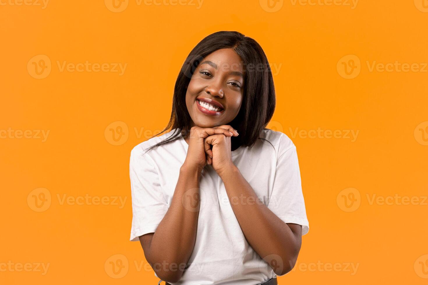 Sweet black lady showing tenderness on orange studio background, touching her chin with both things and smiling at camera, african american woman flirting or seeing something heart melting photo