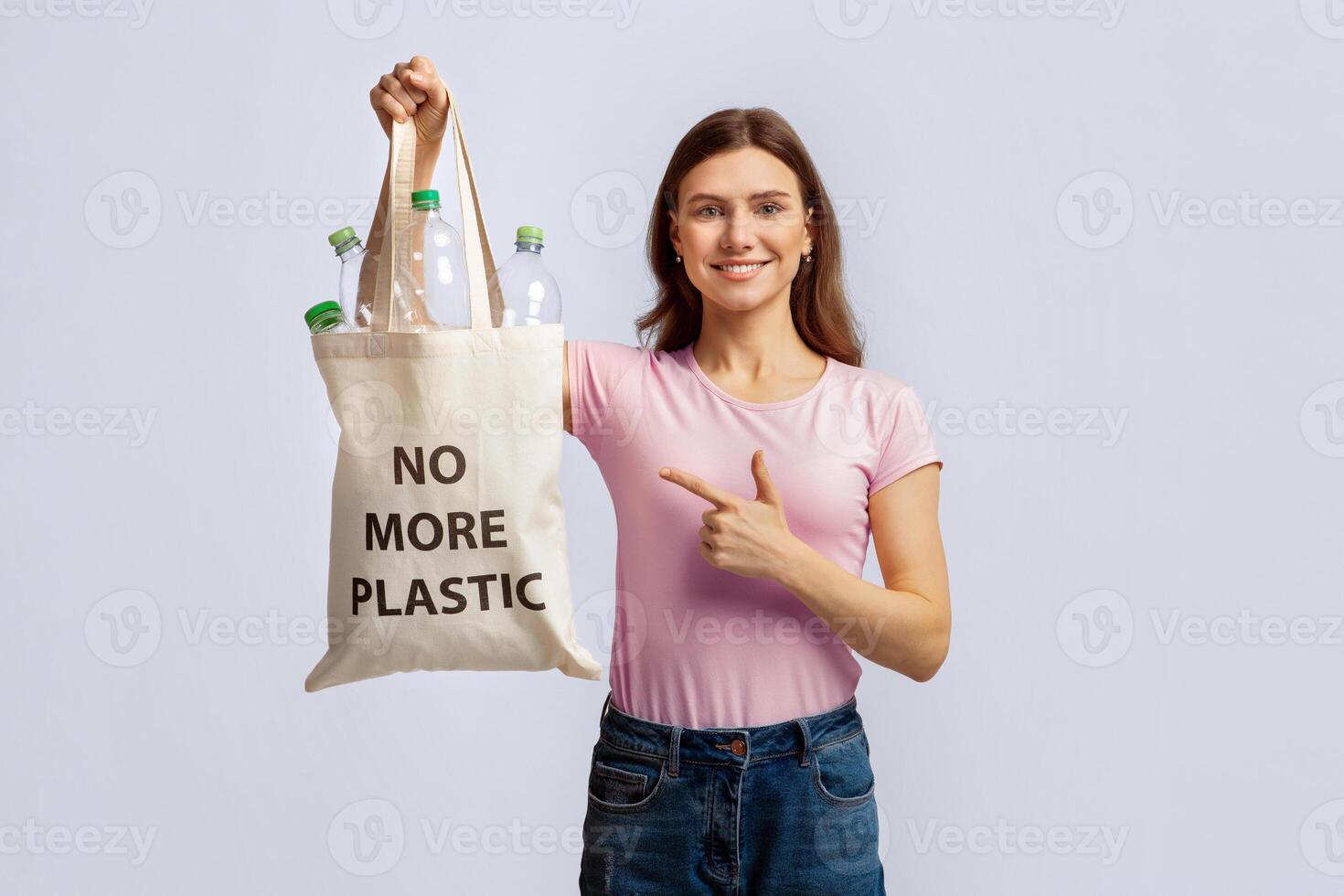 Eco-Activism Concept. Young Smiling Woman Pointing At Tote Bag With No More Plastic Inscription, Collecting Empty Bottles For Reuse And Recycling, Standing Over Grey Studio Background, Copy Space photo
