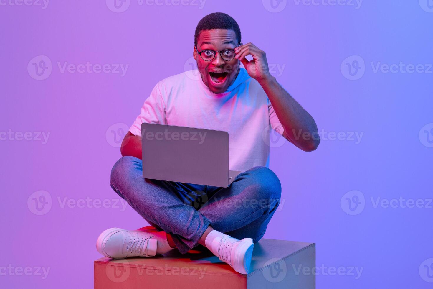Portraif Of Amazed Black Guy Sitting On Cube With Laptop In Neon Purple Light, Looking At Camera With Opened Mouth, Excited African American Male Student Enjoying Modern Technolgies For Education photo