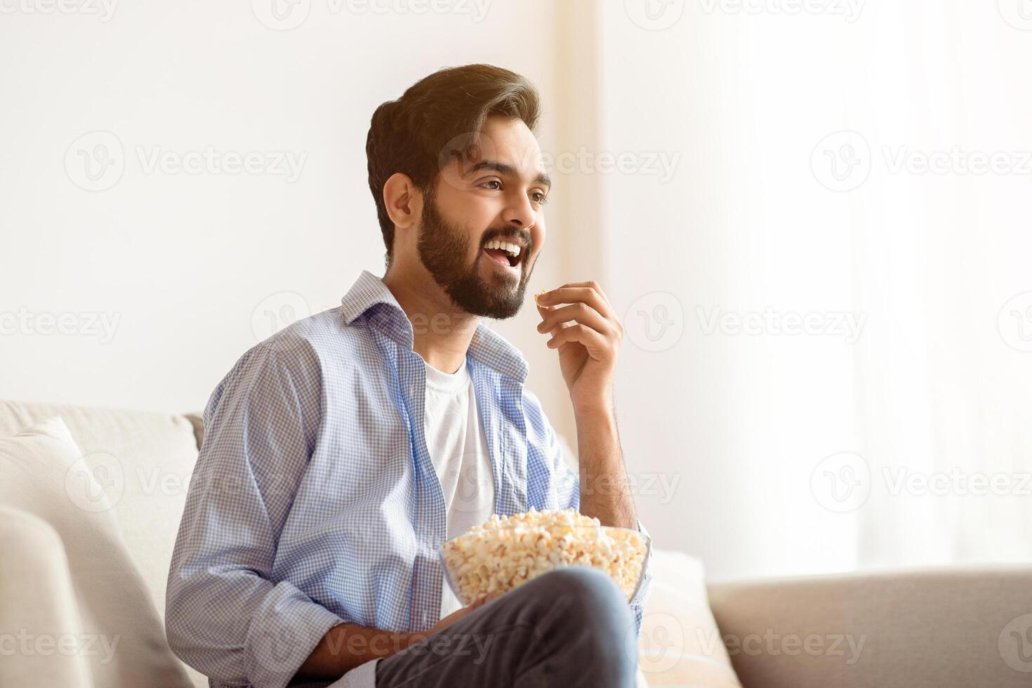 Cheerful Arab Guy Watching Tv And Eating Popcorn At Home, Relaxing On Couch In Living Room, Cheerful Eastern Man Enjoying His Favorite Movie Or Show, Resting On Comfortable Sofa In Casual Atmosphere photo