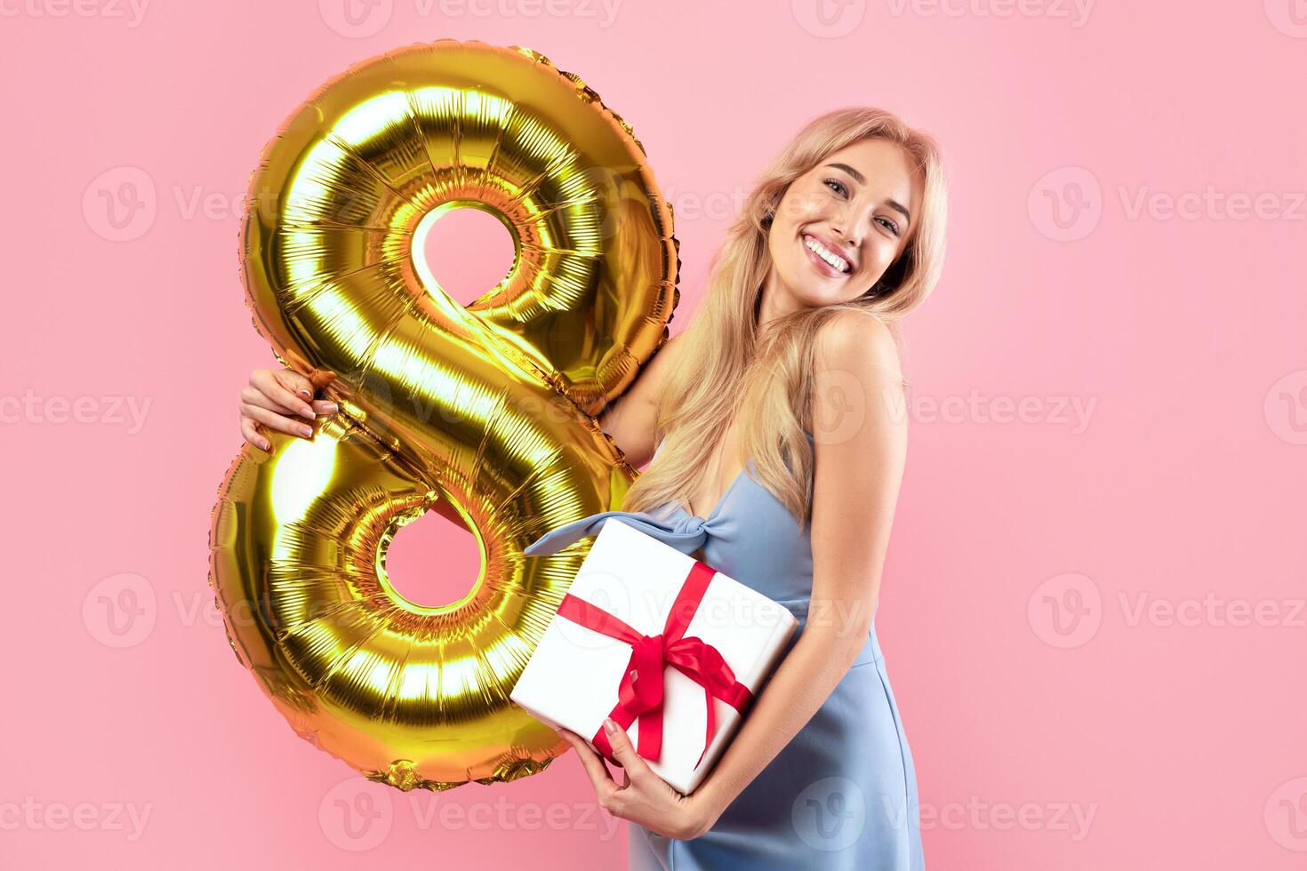 Woman's Day celebration concept. Charming lady with golden eight shaped balloon and gift box looking at camera and smiling over pink studio background, panorama with copy space photo