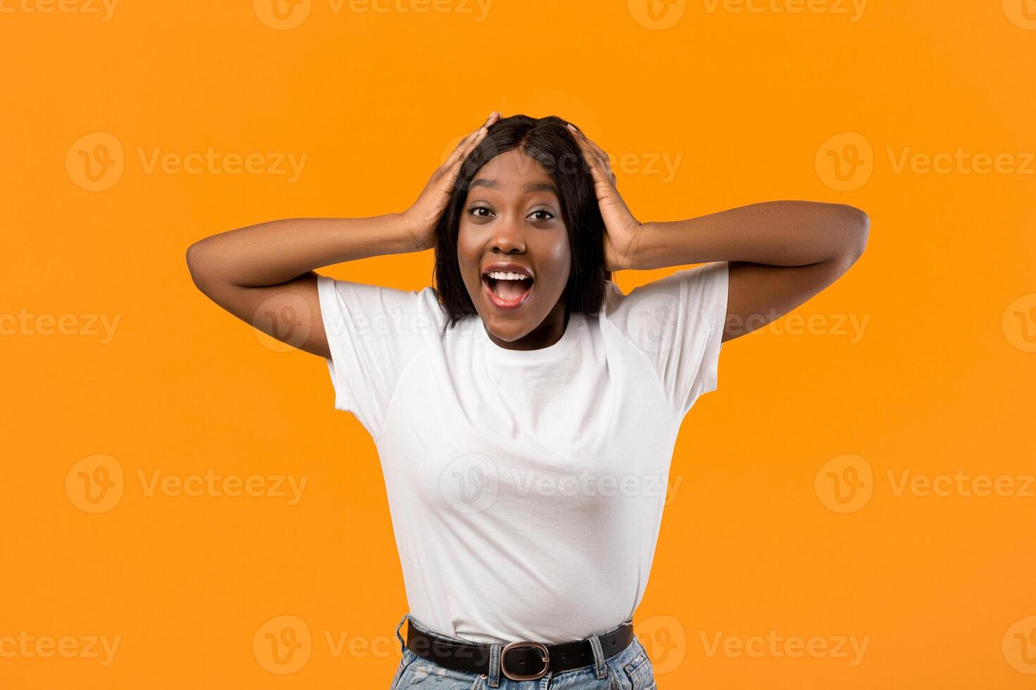 Shocked black lady in white t-shirt raising hands up and touching head over orange studio background, emotional african american young woman expressing amazement, seeing something breathtaking photo