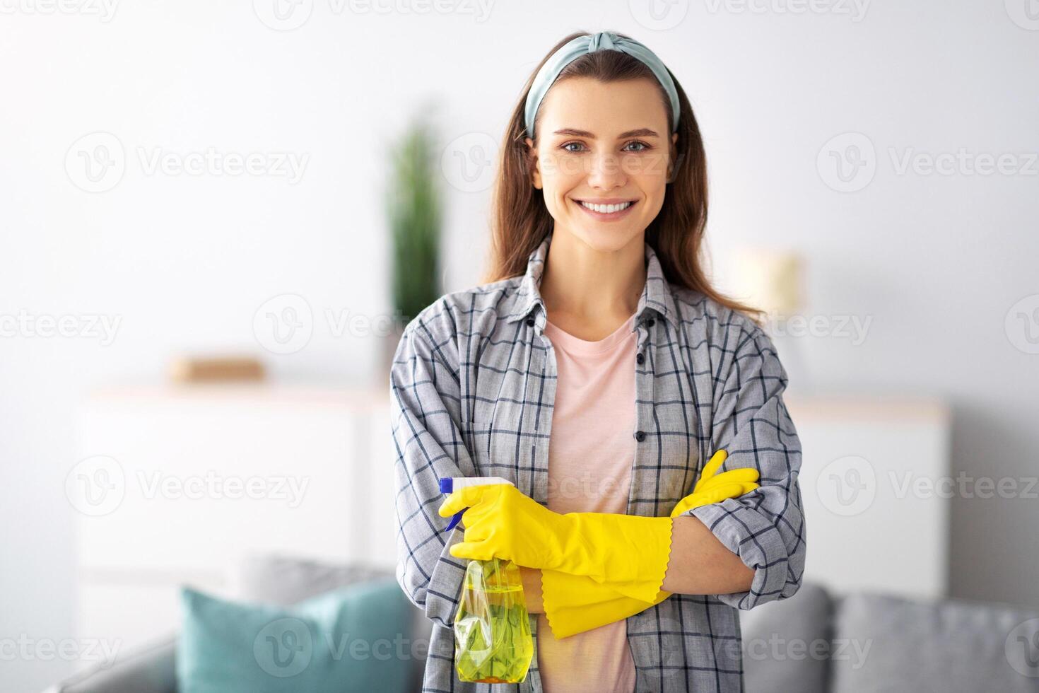 Joyful young housemaid in rubber gloves holding detergent and smiling at camera at home. Lovely millennial lady offering professional housecleaning. Sanitary service concept photo