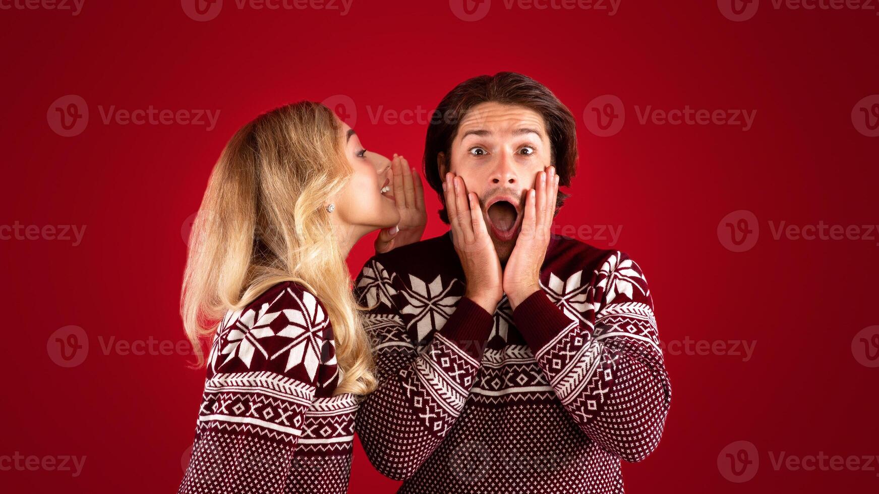 Shocked millennial guy in Christmas outfit and girl whispering something into his ear on red background, panorama photo