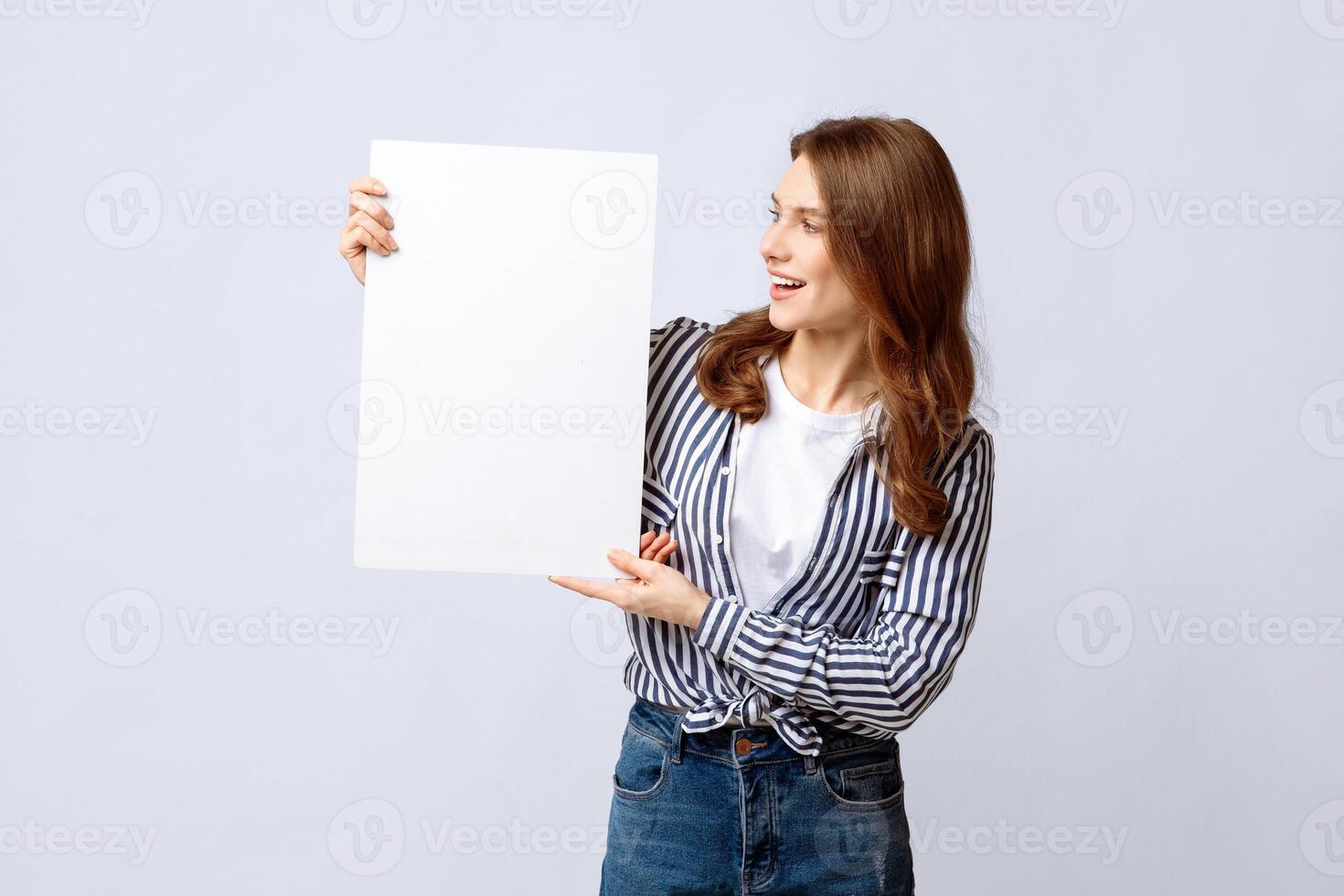 Young Woman Looking With Excitement At Blank White Placard In Her Hands, Millennial Lady Holding Empty Poster With Copy Space For Your Offer Or Advertisement, Standing Over Light Background photo