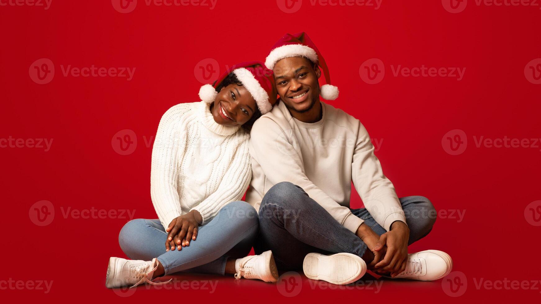Romantic Cristmas. Portrait Of Loving Black Couple Wearing Santa Hats Posing Together While Sitting On Floor Over Orange Studio Background, African Spouses Having Fun During Xmas Holidays, Copy Space photo