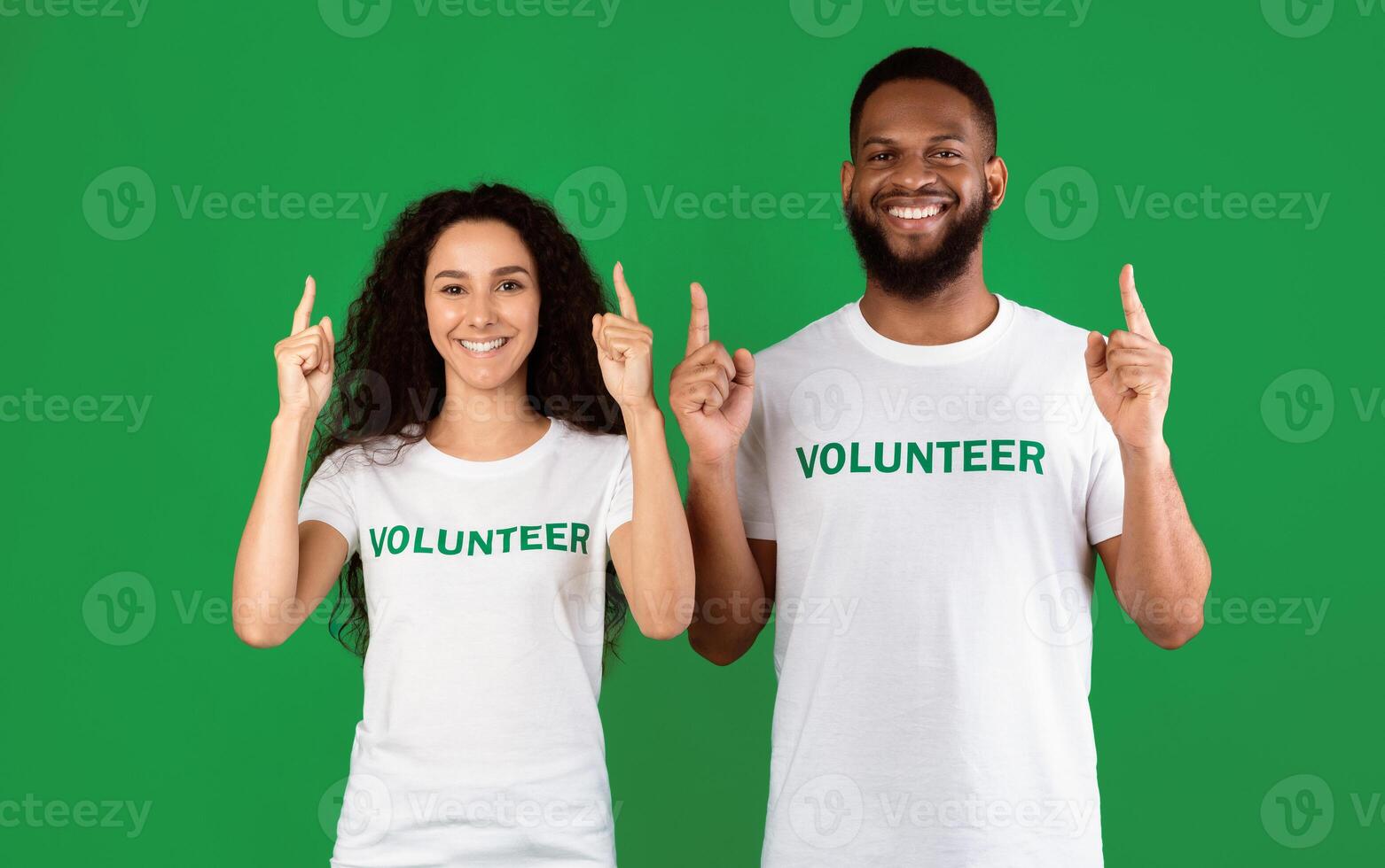 Two Multiracial Volunteers Pointing Fingers Up Advertising Something Standing Over Green Background In Studio, Smiling To Camera. Look There. Charity And Volunteering Ad Banner photo