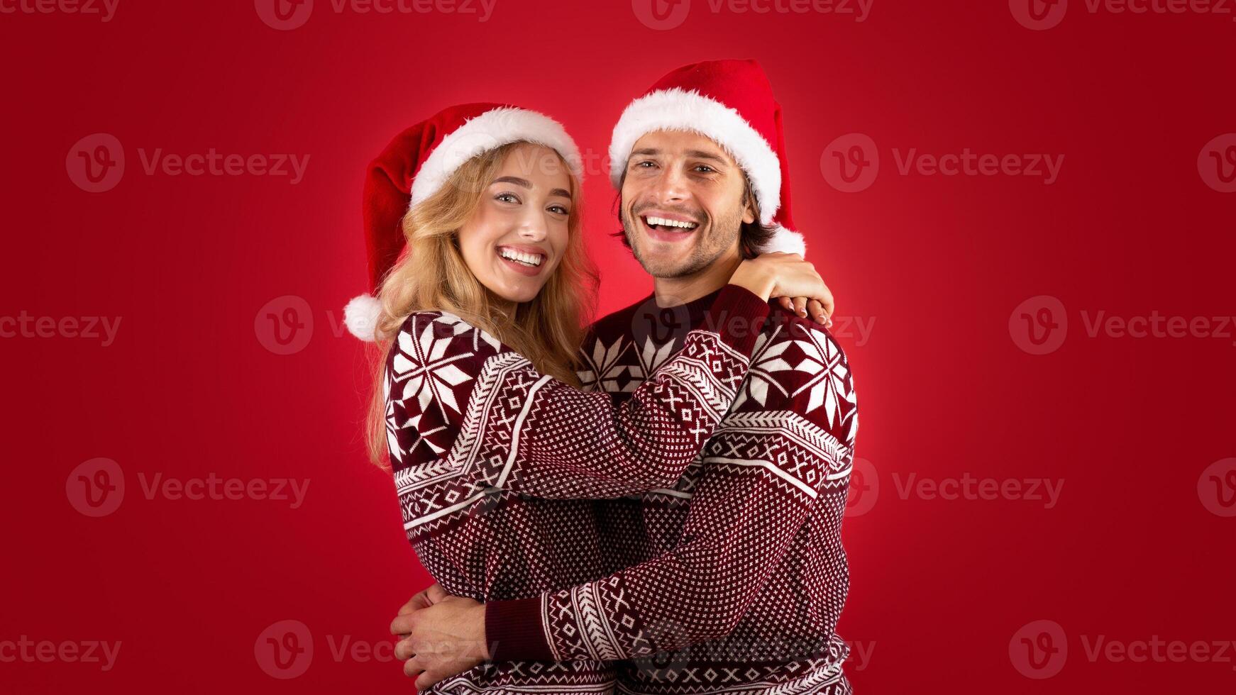 Expressing affection. Lovely young couple in Santa hats and Christmas sweaters hugging on red background photo