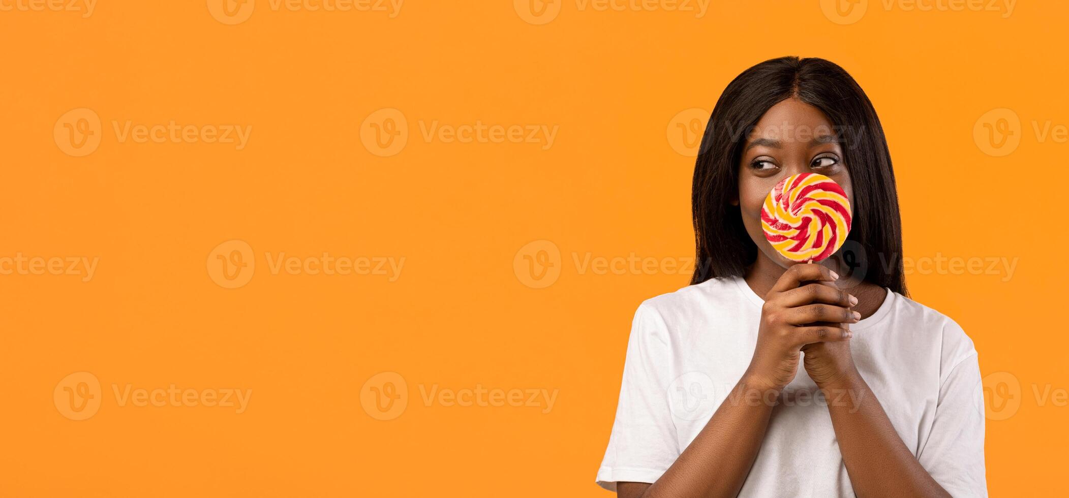 Shy black woman covering her mouth with candy on stick and looking at copy space for advert, orange studio background. Playful african american young lady having fun, holding sweets, panorama photo