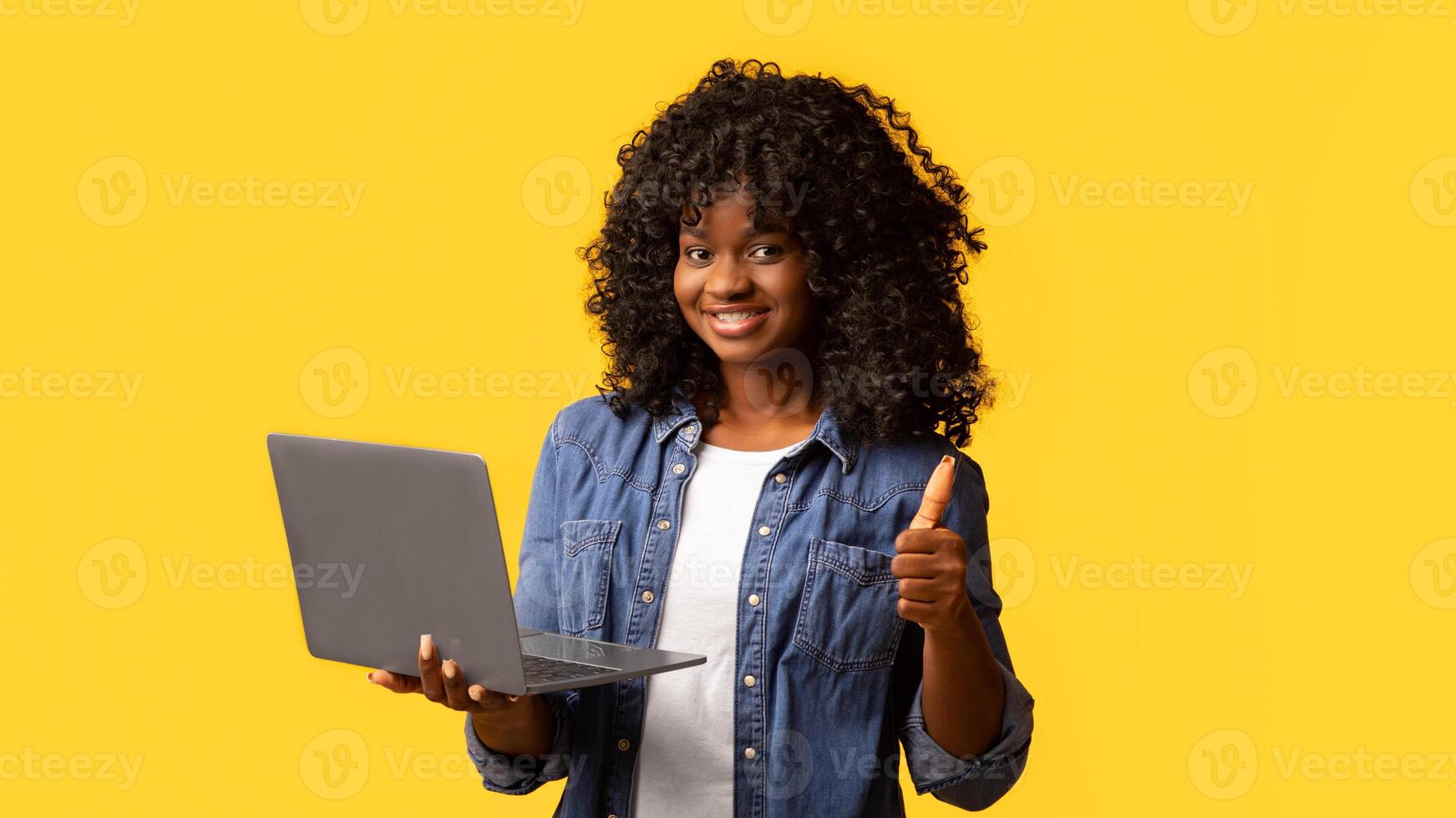 Happy african american young woman holding newest laptop and showing thumb up, yellow studio background, panorama with copy space. Cheerful black lady using notebook, shopping online or websurfing photo