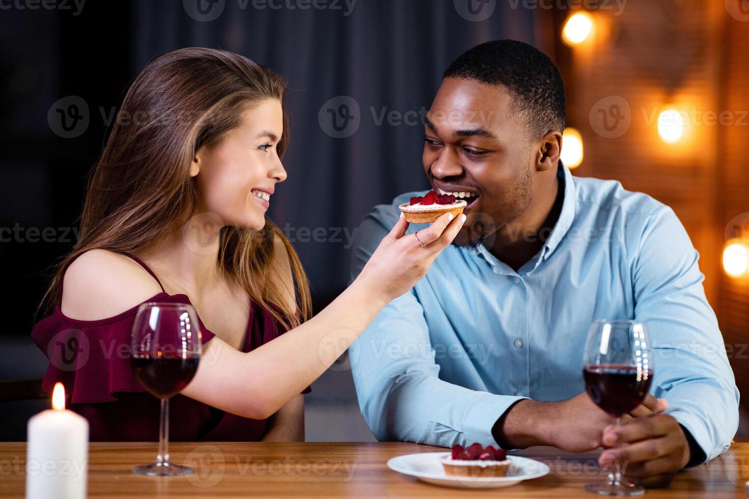 Romantic interracial couple having dinner date in restaurant, feeding each other with desserts and drinking wine, black man and while woman having fun while celebrating valentine's day together photo