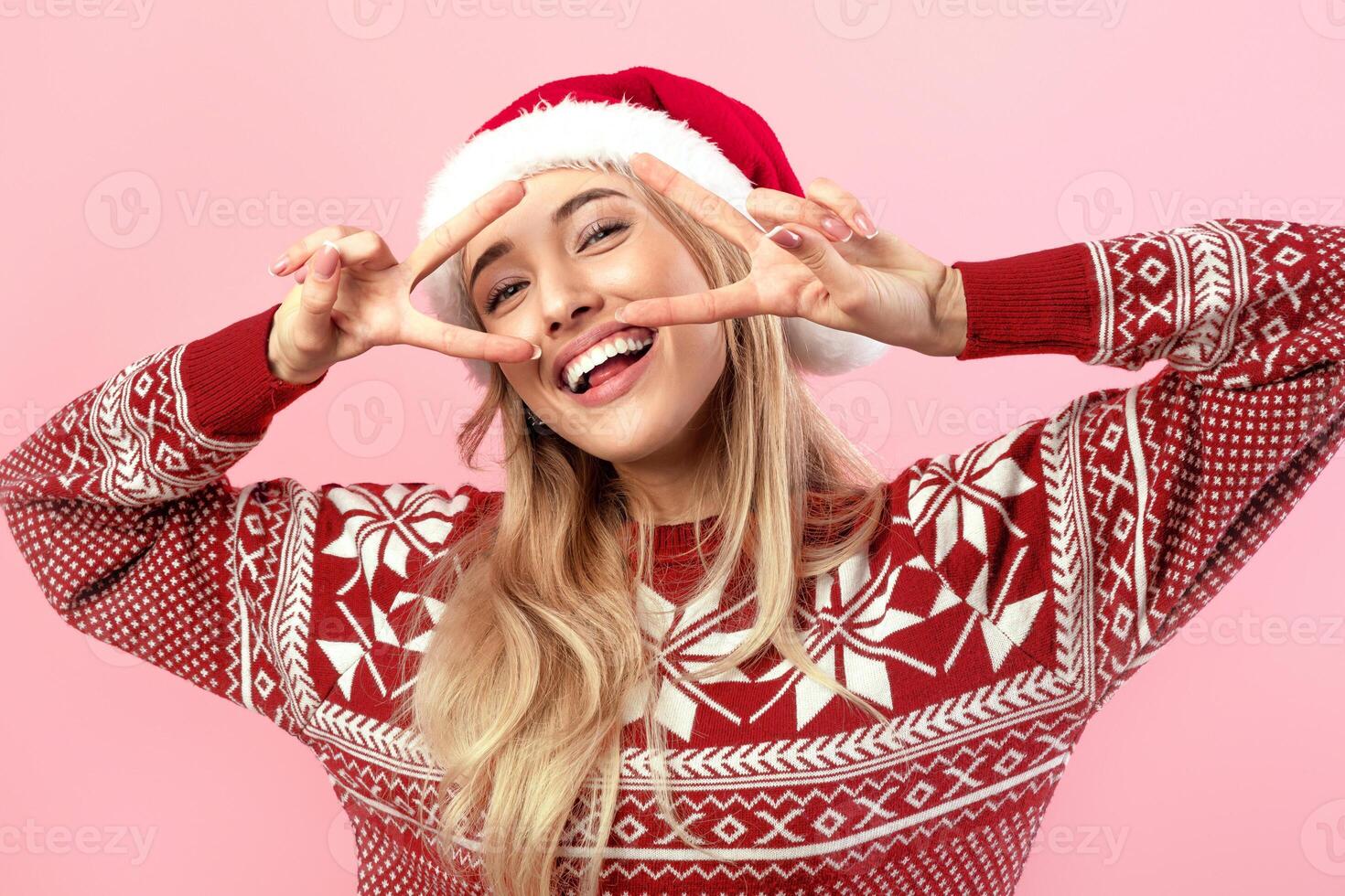 Cheerful young lady in Xmas sweater and Santa hat smiling and making peace gesture with both hands on pink studio background. Pretty millennial woman in winter clothes gesturing victory photo