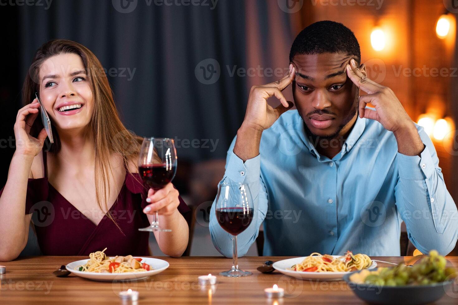 Bored Man Sitting At Date Next To His Drunk Girfriend Talking On Cellphone. Annoyed African American Guy Touching Temples, Feeling Upset And Lonely. Interracial Couple's Problems Concept, Free Space photo