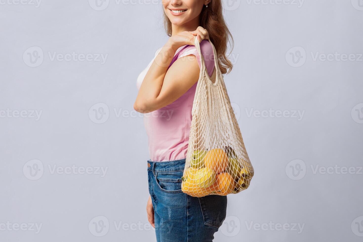 Reducing Waste. Positive Young Lady Using Reusable Eco Bag For Shopping, Unrecognizable Woman Holding Mesh With Organic Fruits And Vegetables, Standing Over Light Studio Background, Copy Space photo