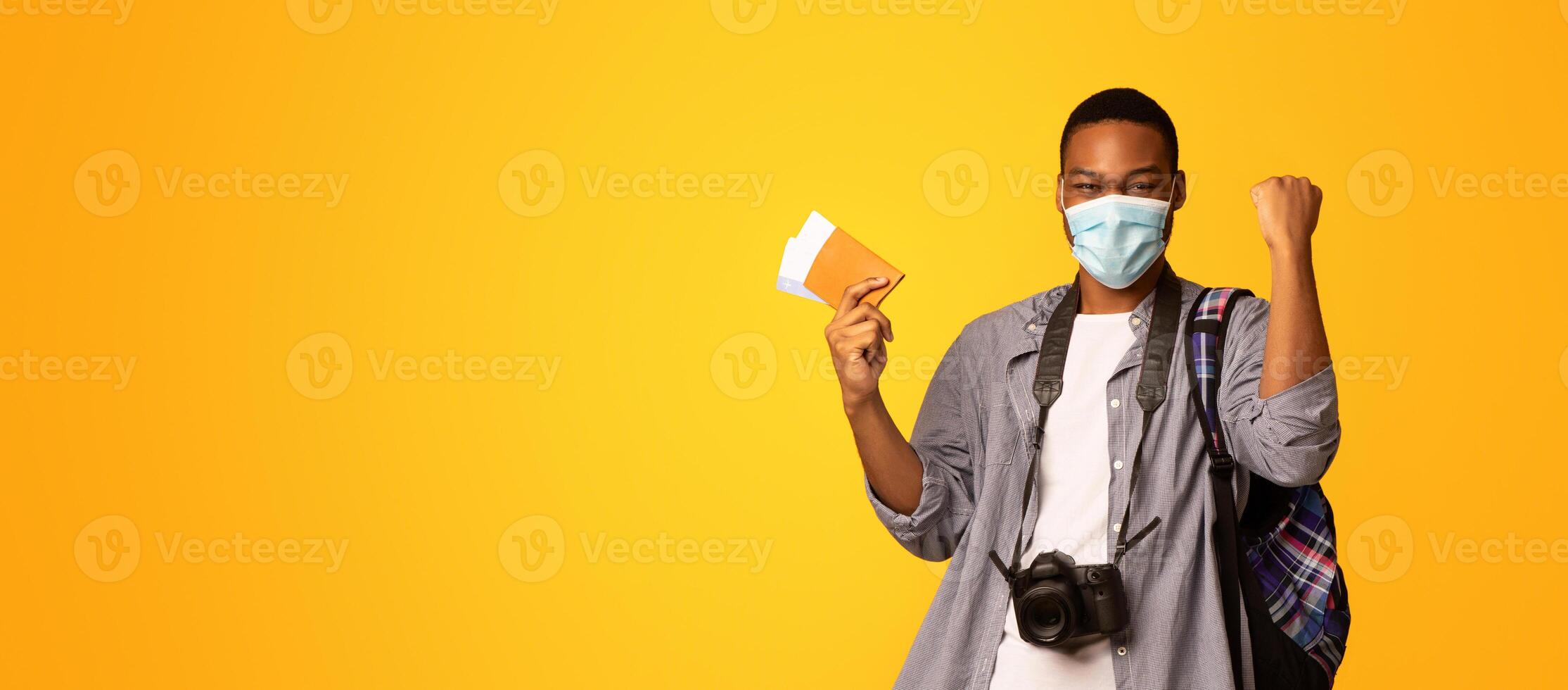 Vacation, Finally. Portrait Of Happy African American Tourist Guy In Medical Face Mask And Passport In Hand Raising Fist In Excitement, Ready For Trip During Coronavirus Pandemic, Yellow Background photo