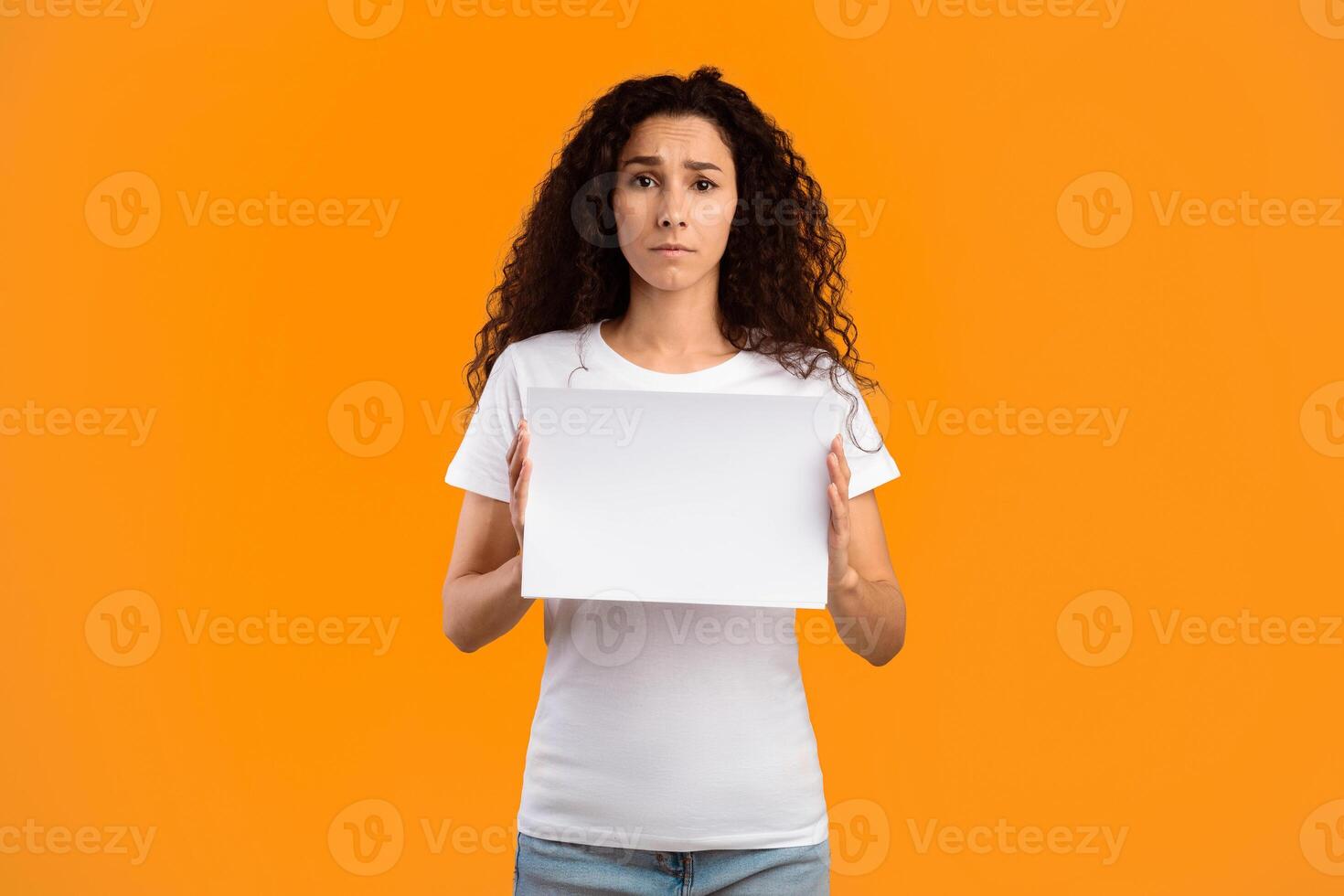 Sad Woman Holding Empty Advertisement Board For Your Text Standing Over Yellow Studio Background, Looking At Camera. Lady Showing White Paper Poster Advertising Something. Mockup photo