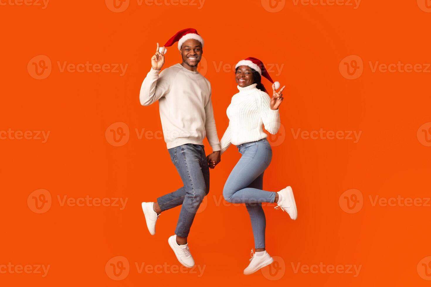 Playful Black Couple In Santa Hats Holding Hands And Showing Peace Gesture, Having Fun While Jumping Together Over Orange Studio Background, Enjoying Celebrating Christmas Holidays, Full Length photo