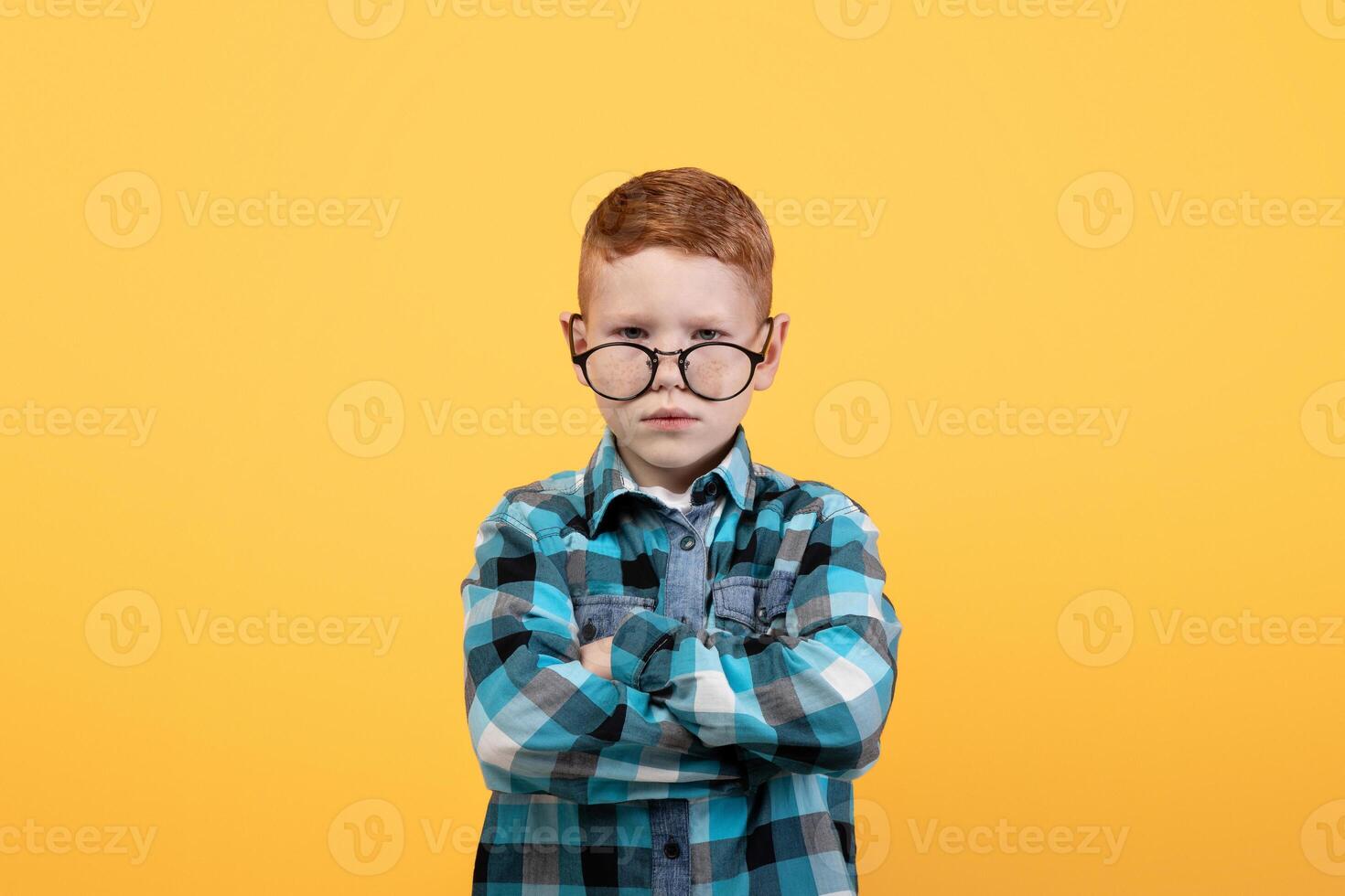 Angry boy in eyeglasses and casual shirt with arms crossed posing over yellow studio background, copy space. Annoyed redhead kid feeling offended, expressing negative emotions photo