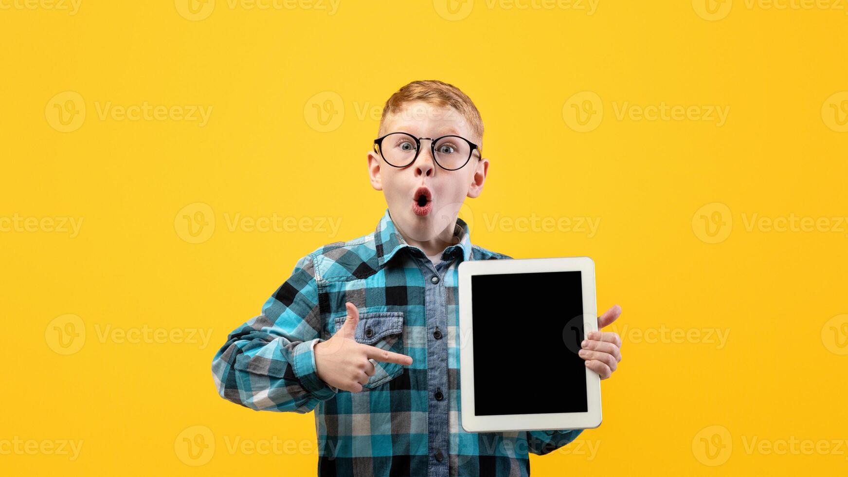 Amazed schoolboy in glasses pointing at empty digital tablet screen, recommending cool educational or entertaining website or application, mockup, yellow studio background, copy space photo