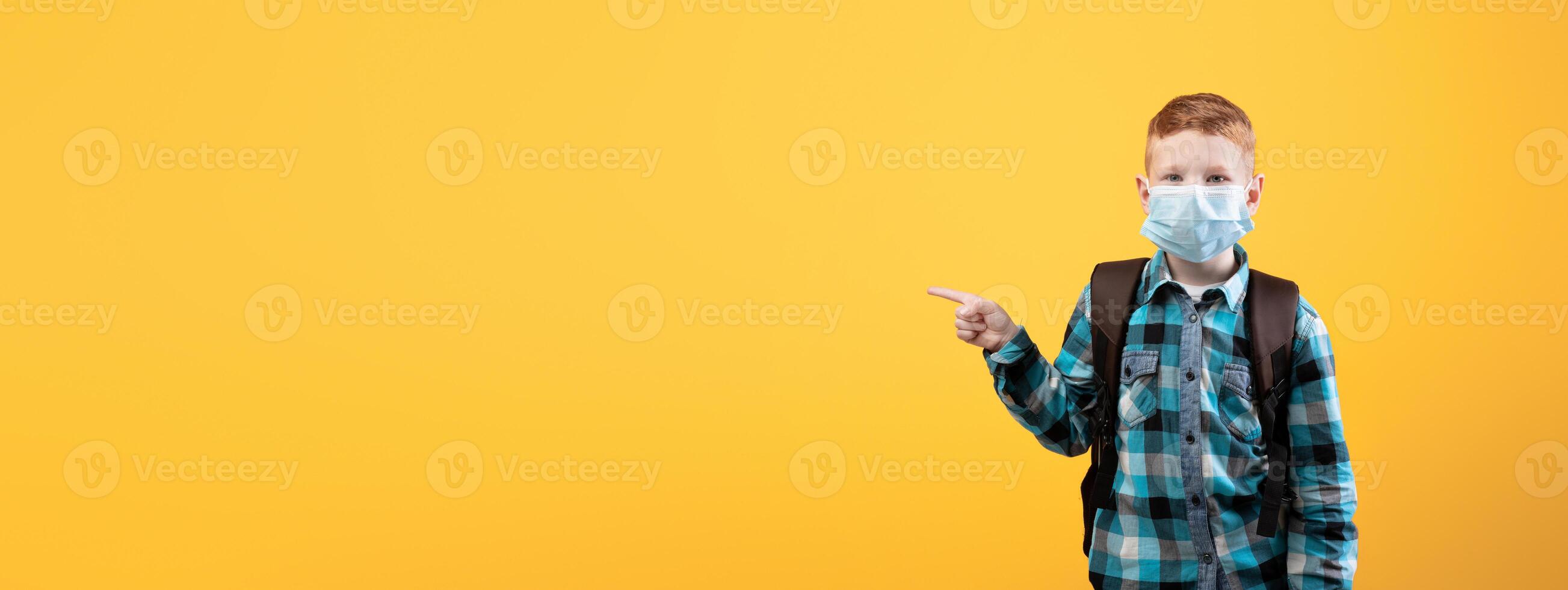 Schoolboy in protective face mask pointing at copy space over yellow studio background. Redhead schoolboy with backpack wearing face mask, using protection against coronavirus while going to school photo