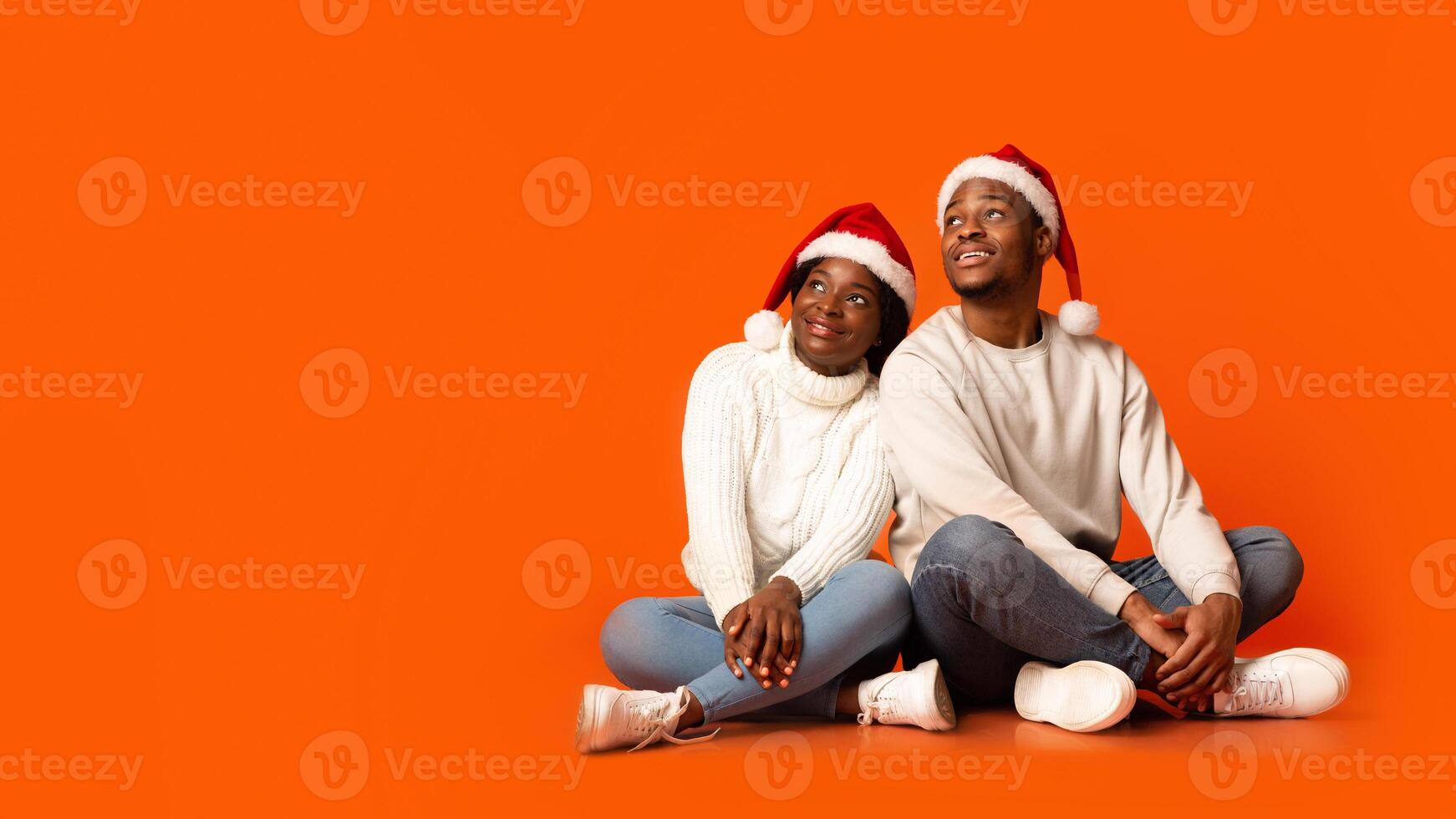 Christmas Promo. Positive Black Spouses In Santa Hats Looking At Copy Space With Interest, Sitting On Floor Over Orange Studio Background, Enjoying Holidays Offers And Xmas Sales, Panorama photo