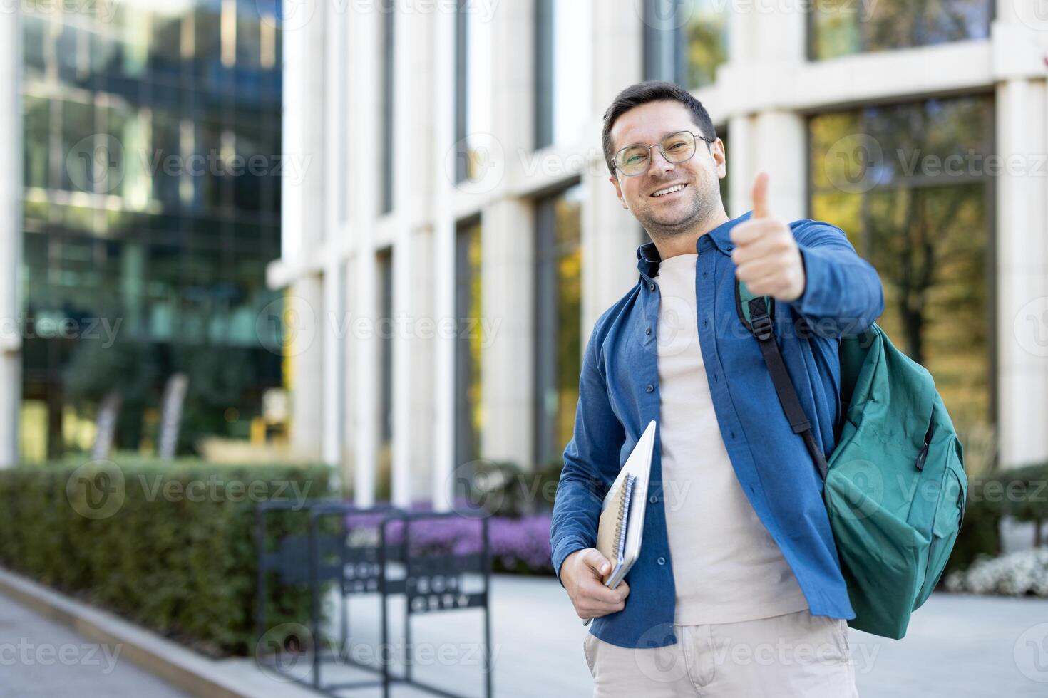 A smiling student with a thumbs-up gesture, wearing a backpack and holding a notebook, stands in front of a modern building. photo