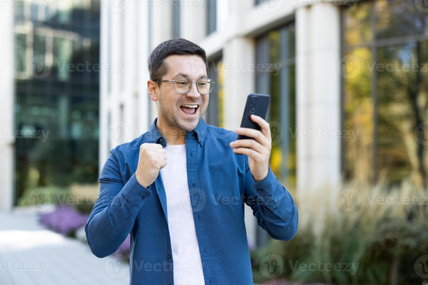 An excited man outside is looking at his phone, celebrating good news. He smiles broadly and raises his fist in joy, demonstrating his accomplishment. photo