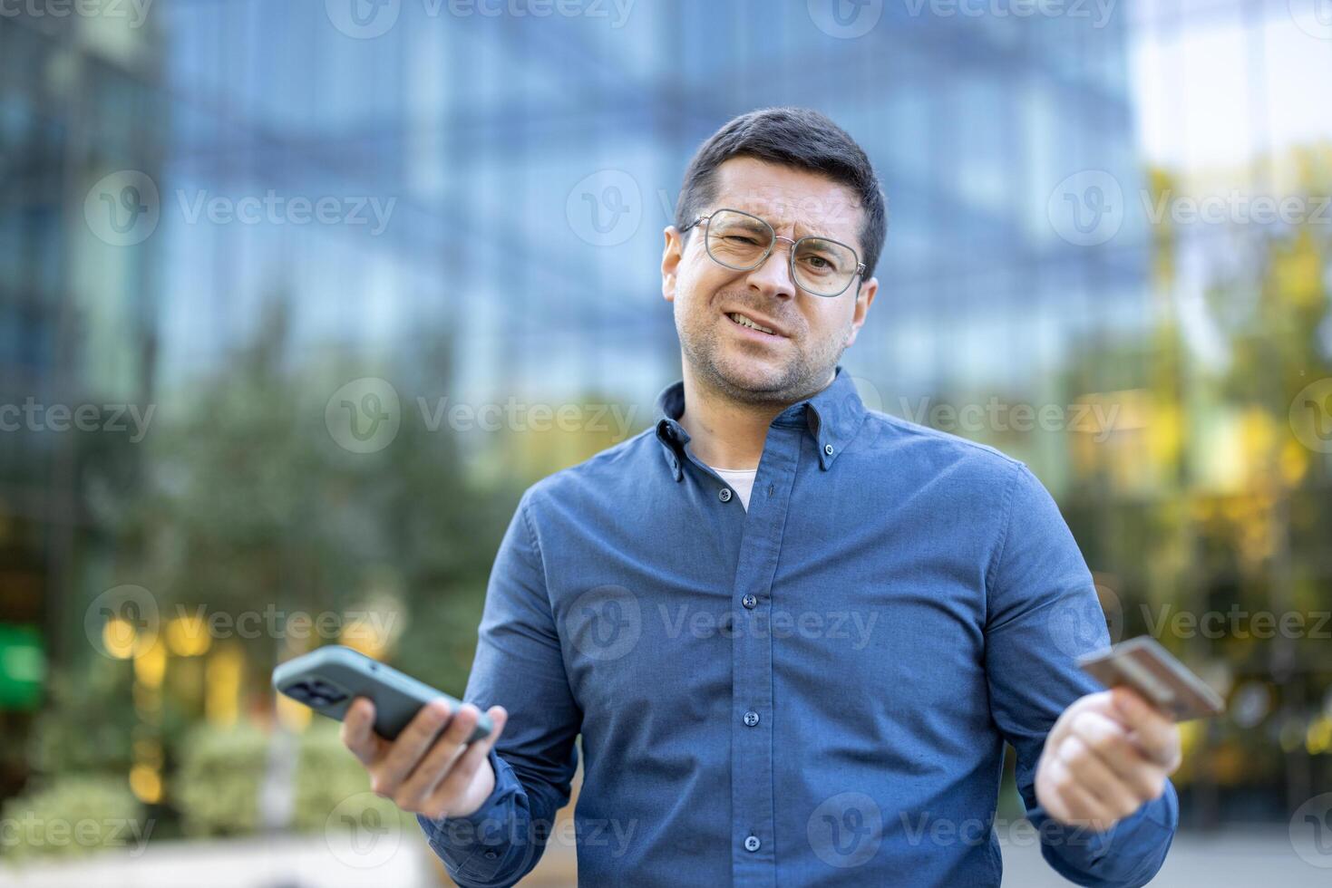 A concerned man outside is holding a phone and a credit card, looking perplexed. This captures financial stress. photo