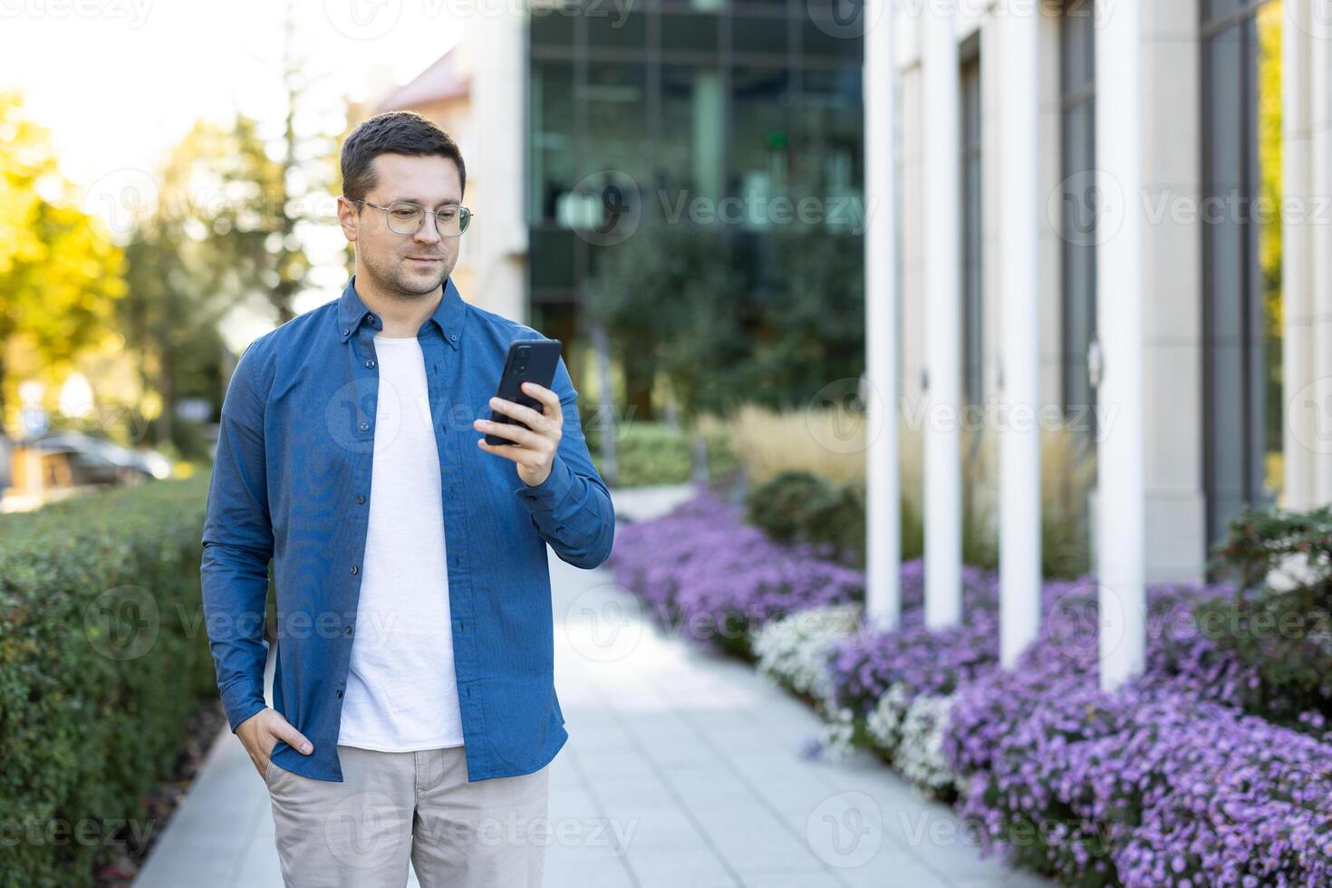 A man in glasses looks at his phone while walking outside in a sunny urban environment, surrounded by flowers and buildings. photo